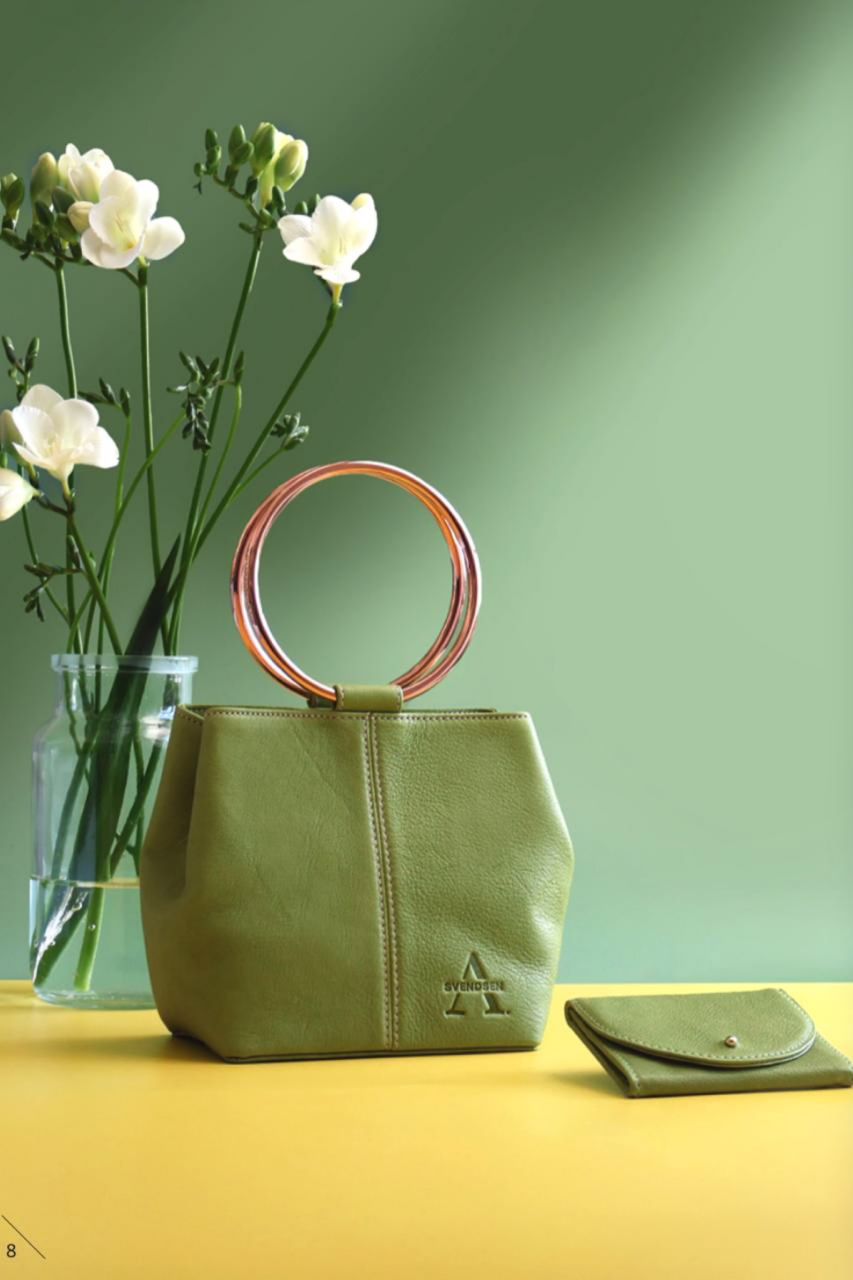 Alexandra Svensen Stillleben einer grünen Ledertasche mit rundem Henkel, arrangiert mit Blumen vor grünem Hintergrund